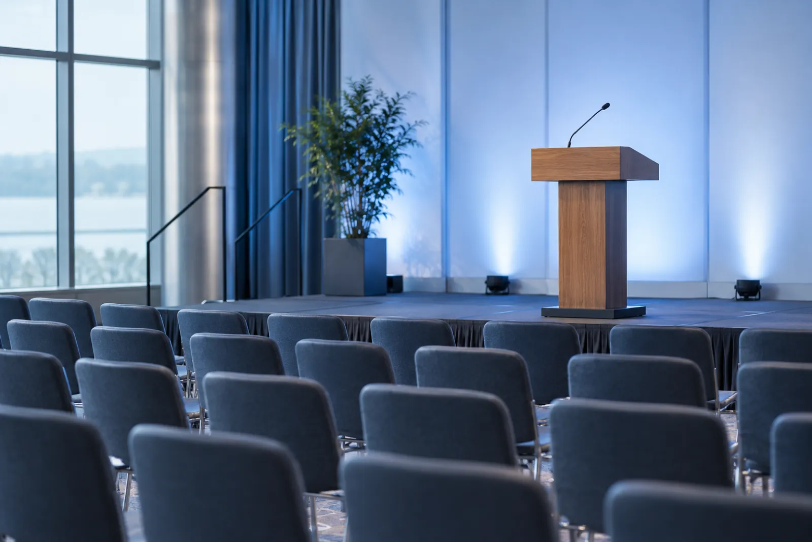 An empty wooden lectern with a gooseneck microphone on a conference stage, rows of empty chairs facing it, cool stage light